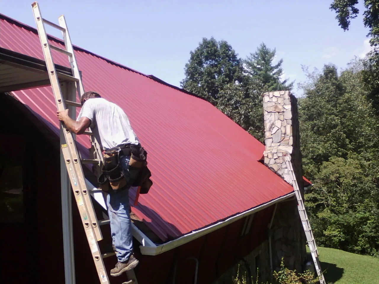 Roofer on a ladder beside a red metal roof and stone chimney — Soffit & Fascia Repair by Roofing Brothers in Edison